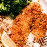 A plate of broccoli, rice, lemon wedge, and breaded tilapia that is flaked with a fork.