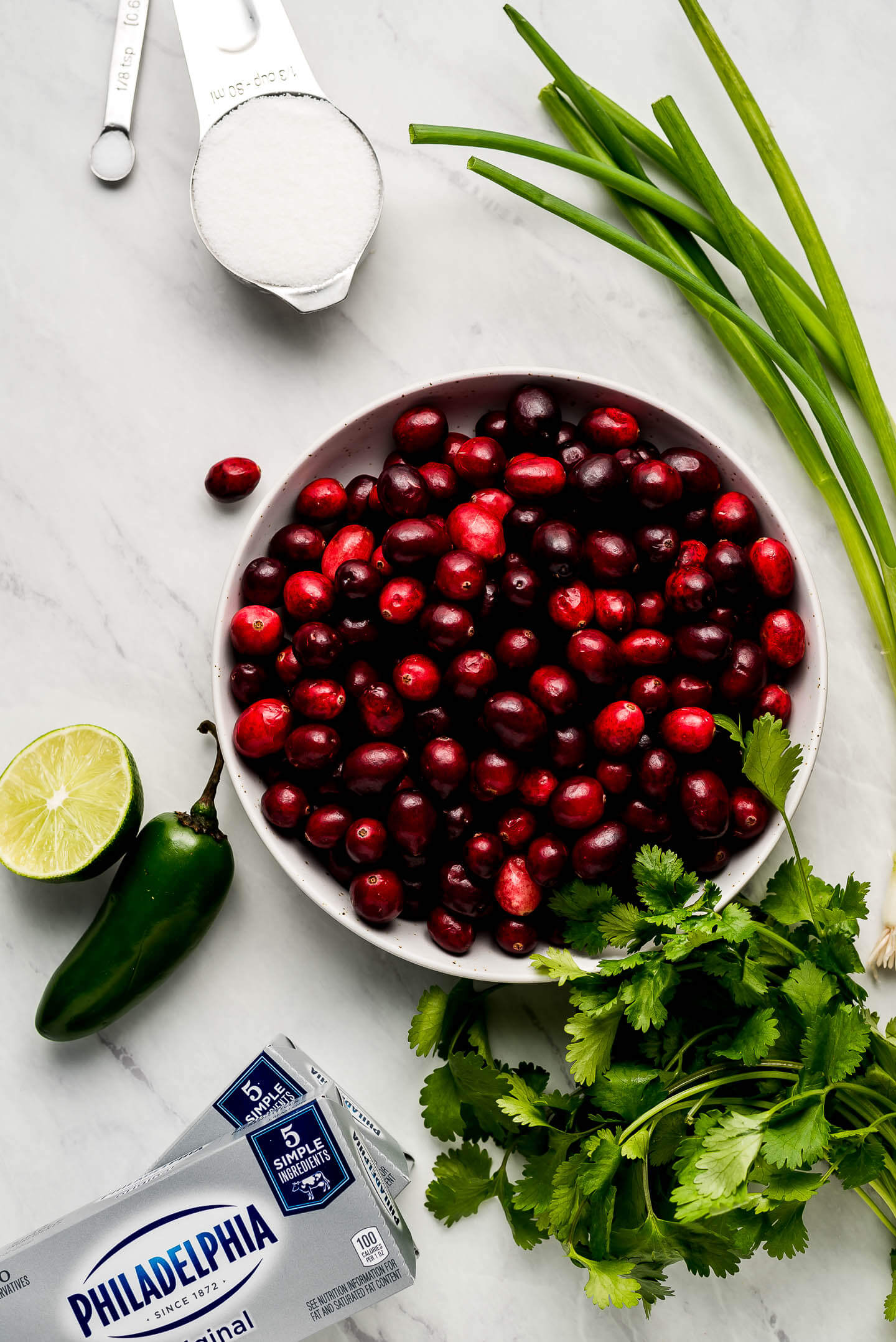 Ingredients on marble surface- bowl or cranberries, sugar, green onions, lime, jalapeno, cilantro, and cream cheese.