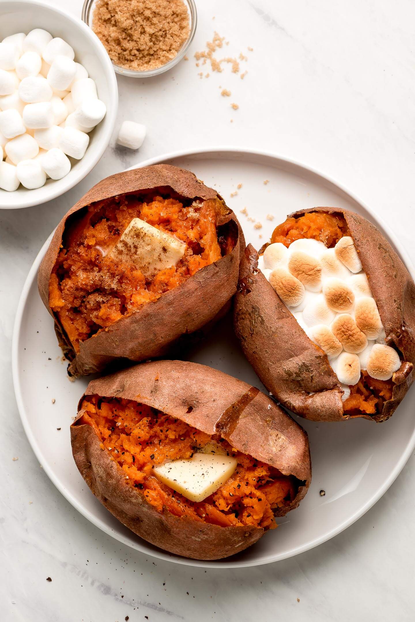 Baked Sweet Potatoes garnished with various toppings and marshmallows and brown sugar to the side.